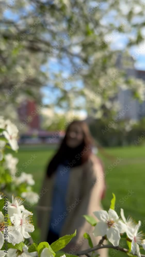 Vertical 4K video. Beautiful young white brunette woman in beige trench coat spins in clearing in city park near flowering tree, bush. Small apple flower. Nature concept. Spring cherry bloom season Vertical 4K video. Beautiful young white brunette woman in beige trench coat spins in clearing in city park near flowering tree, bush. Small apple flower. Nature concept. Spring cherry bloom season