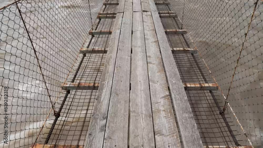 Close-up shot of a fast-flowing brown river running beneath a rope bridge with wooden planks, showcasing a rustic and adventurous setting. Close-up shot of a fast-flowing brown river running beneath a rope bridge with wooden planks, showcasing a rustic and adventurous setting.