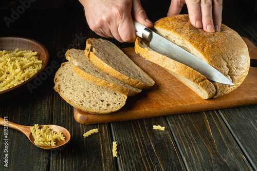 Slicing bread with a knife in the chef hand on a kitchen cutting board before dinner. Low key bakery hearty food concept. Copy space