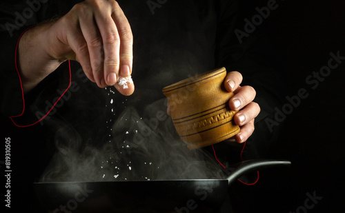 A professional chef adds salt to a hot frying pan while cooking. Black space for advertising.