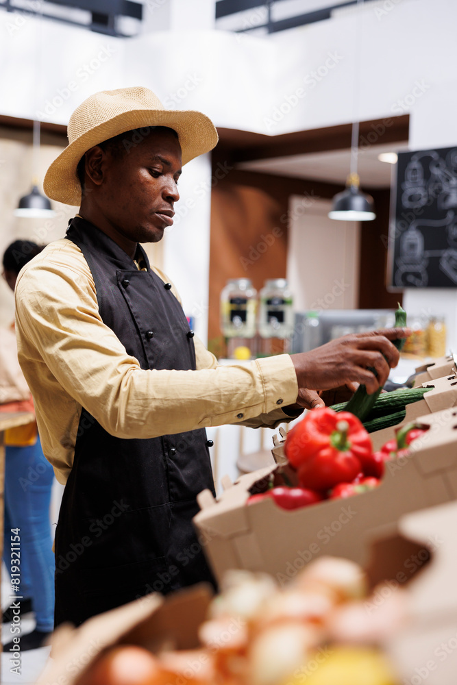 African american man with an apron organizing bio fruits and vegetables in the local supermarket. Selective focus on vibrant red peppers, cucumbers, and onions positioned on shelves. African american man with an apron organizing bio fruits and vegetables in the local supermarket. Selective focus on vibrant red peppers, cucumbers, and onions positioned on shelves.