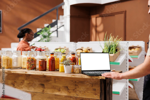 Laptop with white screen is placed on wooden table filled with several glass bottles and jars. In background is a woman looking around the shop while seller uses minicomputer with isolated chromakey.
