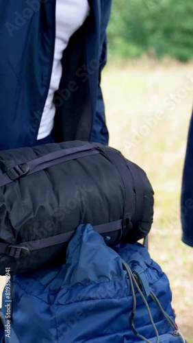 Young man takes thing out of backpack for camping throwing on ground in forest. Black-haired guy wearing blue jacket prepares spot to rest after long hiking Vertical Shot.