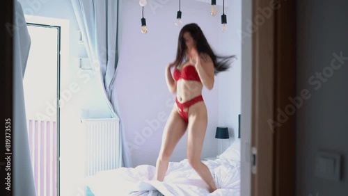 Beautiful brunette woman in red sexy lingerie jumping on bed in modern bedroom