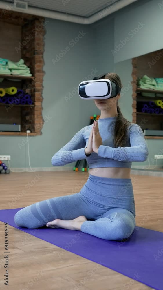Woman in VR glasses sits in Lotus pose with hands in prayer gesture on blue mat in yoga studio. Lady in sportswear enjoys meditating Vertical Shot. Woman in VR glasses sits in Lotus pose with hands in prayer gesture on blue mat in yoga studio. Lady in sportswear enjoys meditating Vertical Shot.