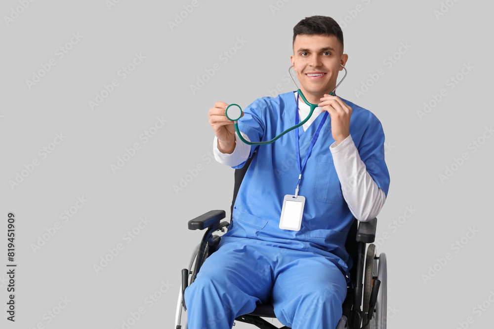 Young male doctor in wheelchair with stethoscope on grey background