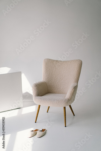 A cozy armchair sits next to a sleek white canvas in a brightly lit studio setting. A pair of casual slip-on shoes rest neatly on the floor  completing this tranquil