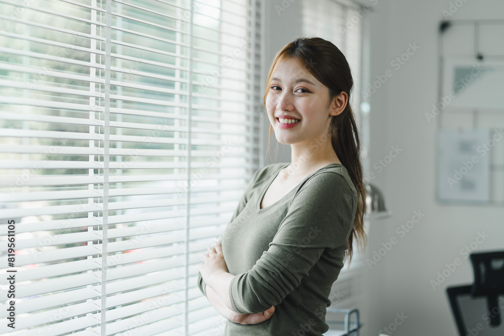 Smiling asian young businesswoman standing by the window in a bright home office. Smiling asian young businesswoman standing by the window in a bright home office.