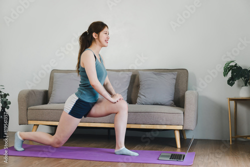 Young Asian woman at home doing yoga in front of open laptop, repeating instructions by professional online fitness trainer.