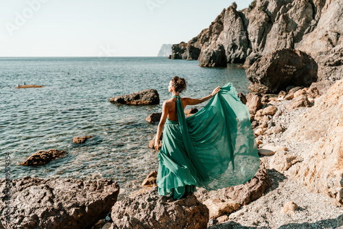 Woman green dress sea. Woman in a long mint dress posing on a beach with rocks on sunny day. Girl on the nature on blue sky background.