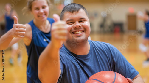 Young adult with down syndrome smiling and giving a thumbs up while holding a basketball in a lively gym environment