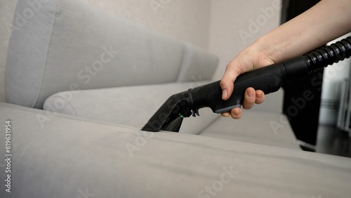 Close-up side view of an unrecognizable woman wearing gloves cleaning a dirty sofa using an upholstery dry cleaning extractor. Close-up: a woman's hand cleans the sofa with a washing vacuum cleaner.