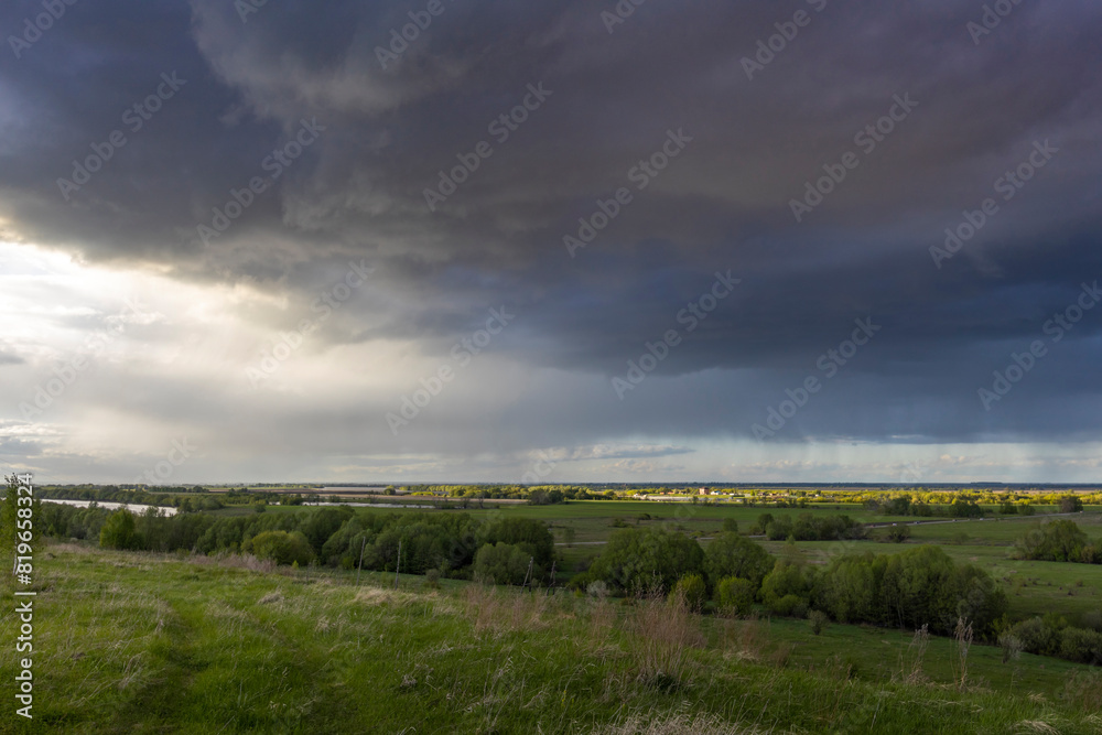 A tranquil rural landscape under a dramatic sky, where storm clouds gather above lush green fields and a distant river, casting a play of light and shadow across the scene