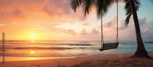 A swing is attached to a palm tree on a sandy tropical beach by the ocean during sunset creating a tranquil scene with a perfect copy space image
