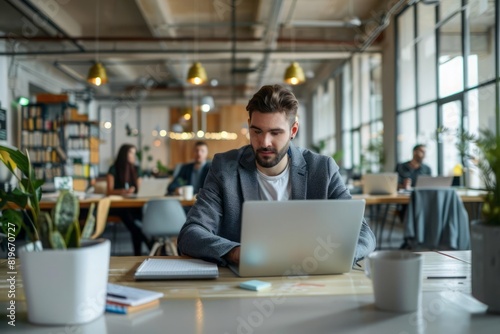 A focused manager researching on his laptop with a view of his office open floor plan and busy colleagues in the background photo