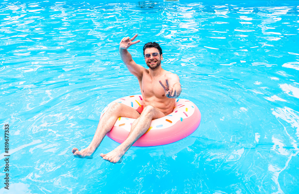 Smiling young man posing at camera and showing victory sign, resting in the swimming pool with inflatable ring. Summertime, holidays, lifestyle concept