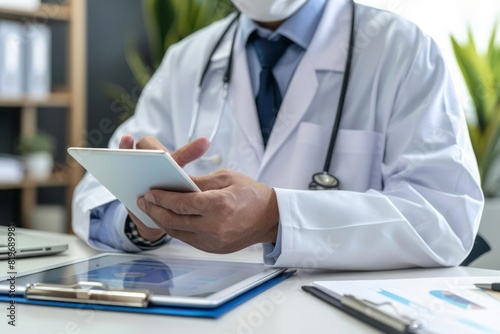 A male doctor sitting at his desk reviewing a patient medical history on a digital tablet before the examination