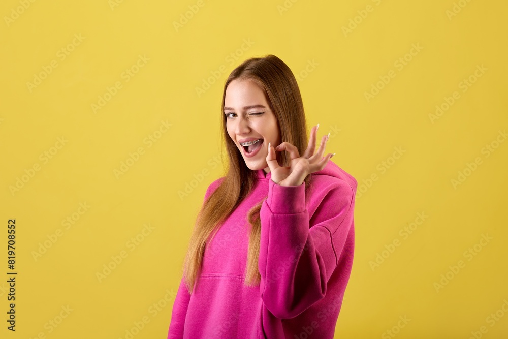 Portrait of a winking young woman making OK sign with fingers and looking at the camera over yellow background. Positive cheerful young woman showing OK sign and winking at camera. Portrait of a winking young woman making OK sign with fingers and looking at the camera over yellow background. Positive cheerful young woman showing OK sign and winking at camera.