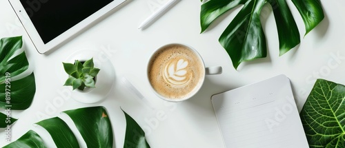 Neat flat lay of a notepad, coffee cup, tablet, and green leaves on a white background, highlighting productivity and simplicity