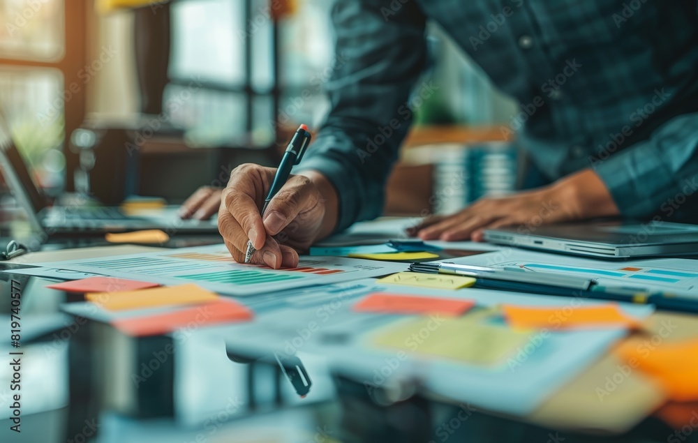 Person working on financial documents at a desk filled with charts and colorful sticky notes in a modern office Person working on financial documents at a desk filled with charts and colorful sticky notes in a modern office