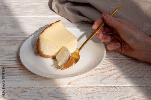 Woman hand cutting tasty piece of cheesecake New York with a golden fork on a wooden table.
