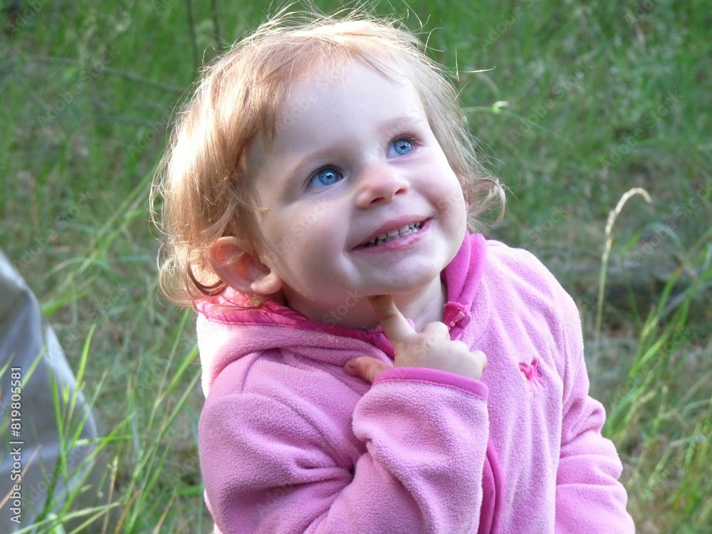 A child with a pink jacket stands on grass and looks up with a curious and inquisitive expression A child with a pink jacket stands on grass and looks up with a curious and inquisitive expression