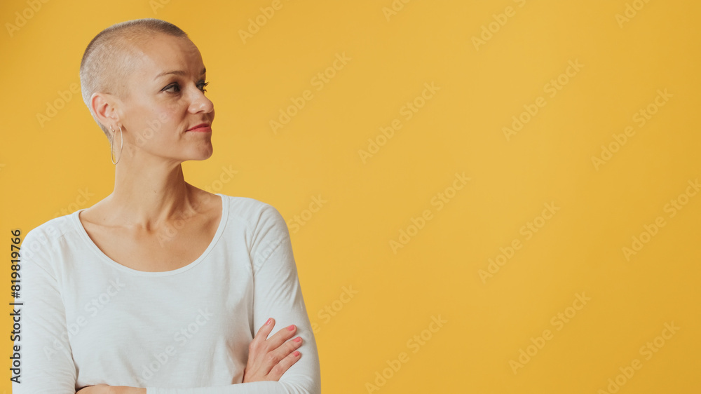 Young hairless woman, with arms crossed and smiling, looking to side at mockup space isolated on yellow background in studio Young hairless woman, with arms crossed and smiling, looking to side at mockup space isolated on yellow background in studio
