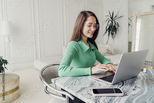 A professional woman with glasses, wearing a green sweater, working on a laptop at a stylish desk.