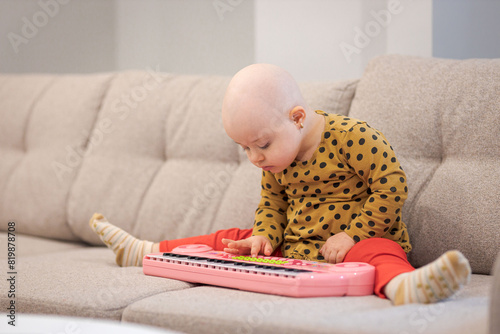 Baby girl with cancer and hair loss due to chemotherapy playing with a piano on sofa © bymandesigns