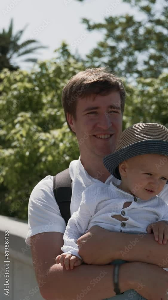 Man holds toddler boy in arms standing on observation deck against lush trees. Father and son enjoy beautiful views on summer vacation in seaside town Vertical Shot. Man holds toddler boy in arms standing on observation deck against lush trees. Father and son enjoy beautiful views on summer vacation in seaside town Vertical Shot.