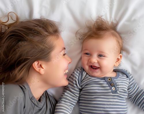 A happy baby lying on the bed with his mother, laughing and looking at each other against a white background. 