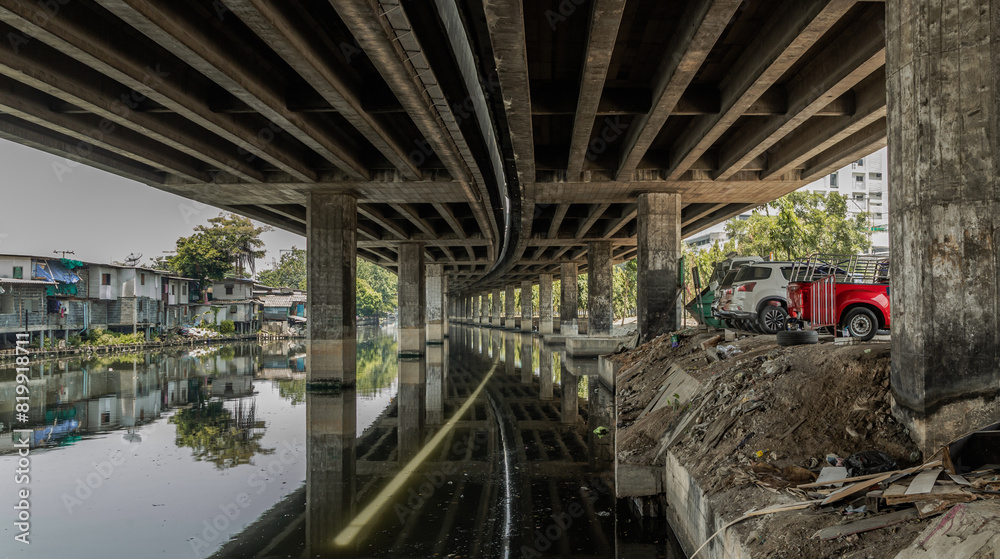 Perspective view of concrete pillars under Khlong Toei Expressway with Car parking and houses are old along the Khlong Phra Khanong. Copy space, Selective focus. Perspective view of concrete pillars under Khlong Toei Expressway with Car parking and houses are old along the Khlong Phra Khanong. Copy space, Selective focus.