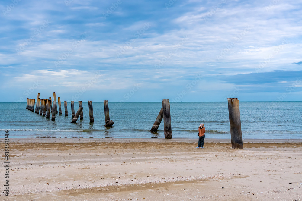 Young Islamic woman using mobile phone with beautiful view of many old leaning pillars of the old pier at slanted concrete pole beach. The joy of spending an outdoor holiday with beautiful nature. Young Islamic woman using mobile phone with beautiful view of many old leaning pillars of the old pier at slanted concrete pole beach. The joy of spending an outdoor holiday with beautiful nature.