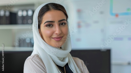 A woman with a hijab smiling at the camera in an office setting with shelves and a whiteboard in the background.