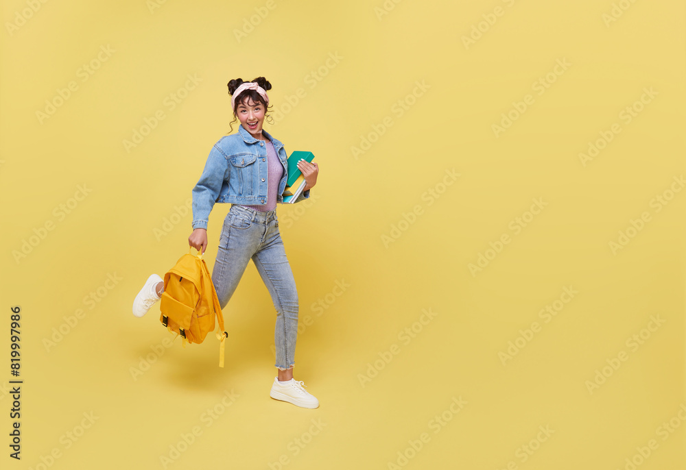 Fototapeta premium Portrait of happy smiling young Asian college student with notebook and backpack walking ready to school isolated on yellow background.