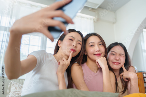 Three young Asian women taking a selfie with mobile phone on a couch in a bright room, showcasing a fun and cheerful moment with playful expressions.