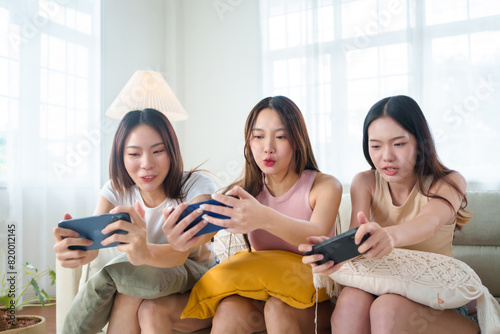 Three young Asian women focused on playing mobile games together on a couch, illustrating concentration and friendly competition in a bright room.