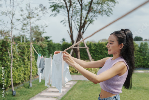 Asian Woman smiling while hanging clothes on a clothesline in a sunny backyard, capturing a moment of contentment during a daily chore.
