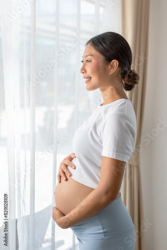 Smiling pregnant woman holding belly, standing by window in bright room. Peaceful and content moment during pregnancy. Free Download Smiling pregnant woman holding belly, standing by window in bright room. Peaceful and content moment during pregnancy. - GFXLoad