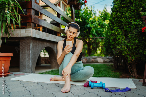 One young caucasian woman is sitting on yoga mat with wireless headphones and mobile phone preparing for training 