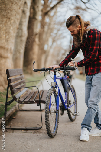 A bearded man in a plaid shirt attentively adjusts the handlebar of a mountain bike next to a park bench.