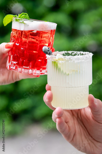 Two hands holding glasses of colorful beverages, garnished with mint and a berry, suggesting a festive or social occasion