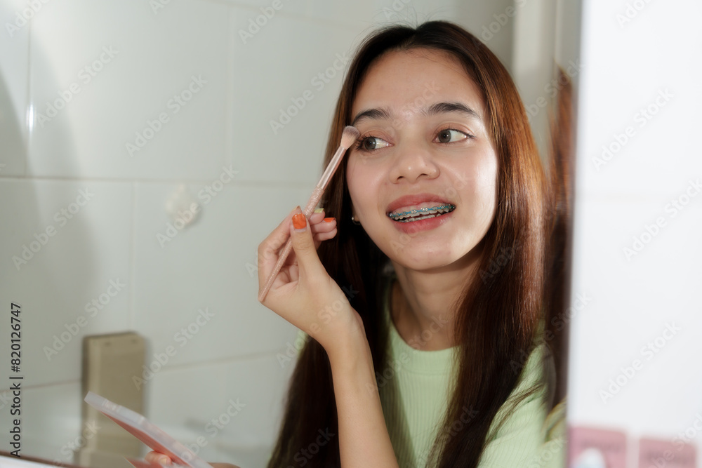 Asian teenage girl with long brown hair, wearing light green sweater, applying eyeshadow with brush, smiling, and showing braces. Indoor happy and fresh expression, emphasizing makeup routine. Asian teenage girl with long brown hair, wearing light green sweater, applying eyeshadow with brush, smiling, and showing braces. Indoor happy and fresh expression, emphasizing makeup routine.