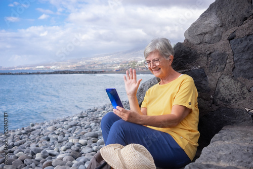 Attractive senior woman sitting on a pebble beach at sea using mobile phone in video call. Mature woman smiles holding cellphone waving hand. Concept of vacation, freedom and happy retirement