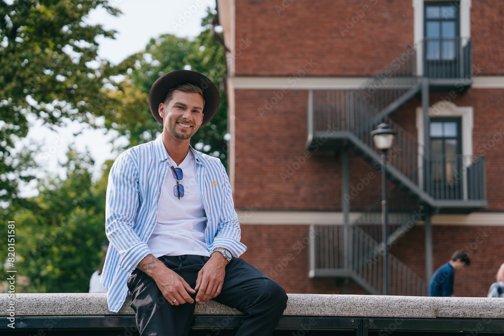 Fototapeta premium man in a striped shirt and hat sits on a ledge, smiling, in a park with a red brick building in the background