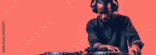 The photo shows a man wearing headphones and mixing music on a turntable. He is wearing a casual outfit and has a focused expression on his face. The background is a solid color. photo