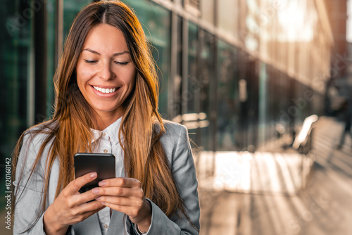 Portrait of beautiful business woman standing in front of her office building and smiling while reading text on her mobile phone.