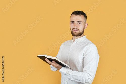 Billede på lærred A young man with a beard in a white shirt on a yellow background holds a large book similar to the Bible and preaches