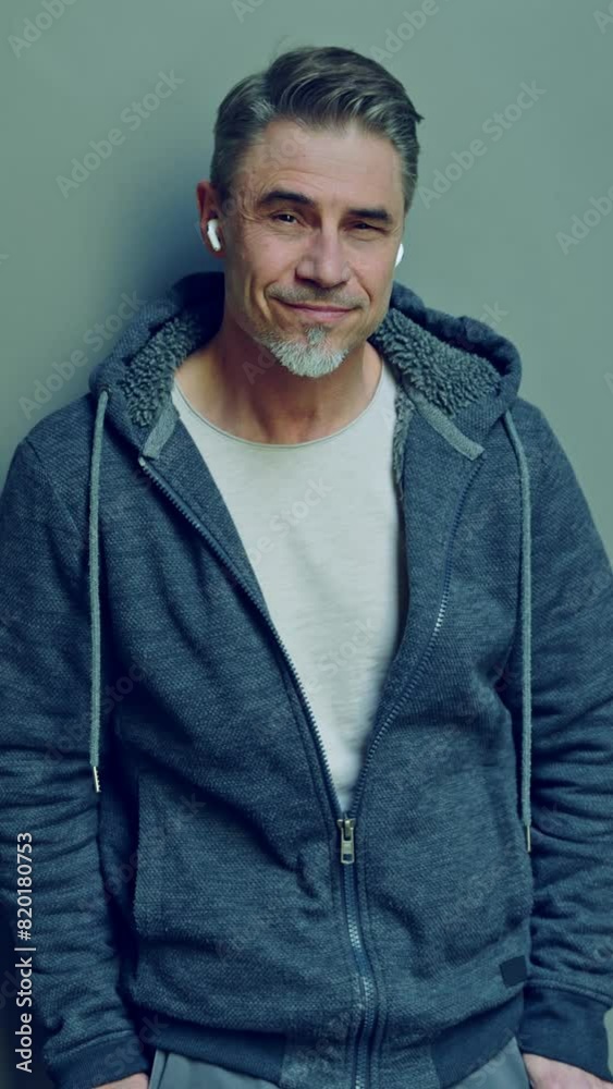 Portrait of a smiling mature man in casual wear with a gray backdrop. Happy mid adult male in 50s, bearded, grey hair, smiling, listening music or podcast in earbuds.