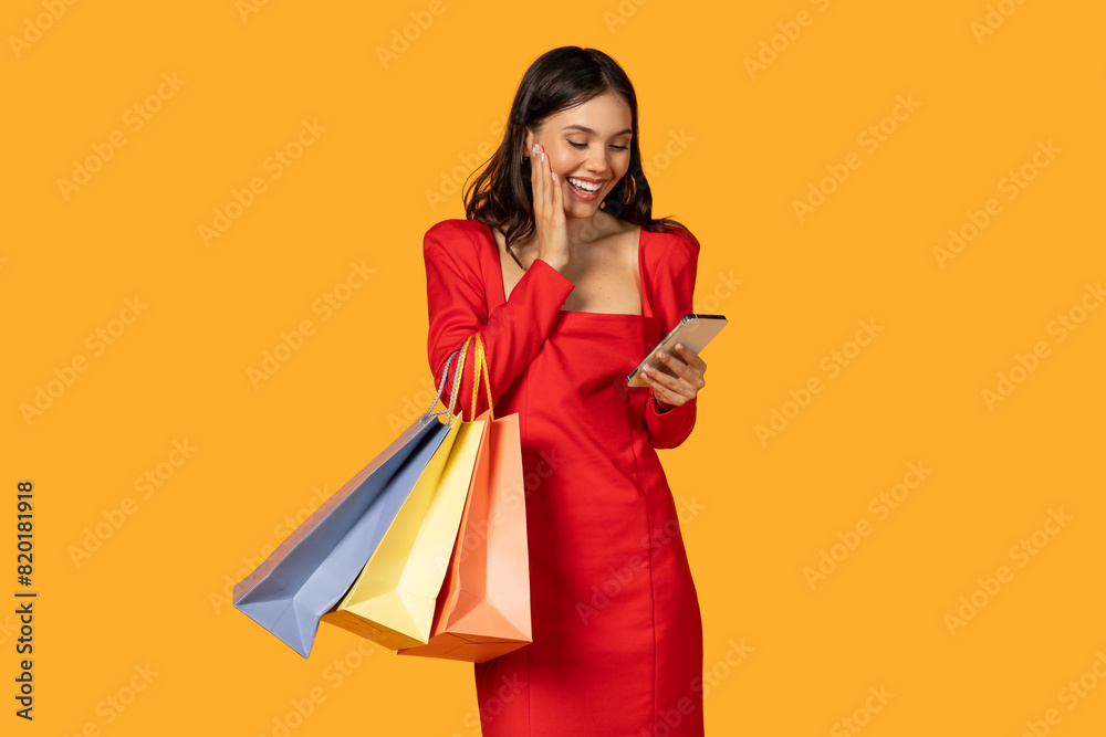 A woman in a vibrant red dress is preoccupied with her smartphone while holding multiple shopping bags in her hands. She seems engrossed in her device, potentially checking messages or making plans.